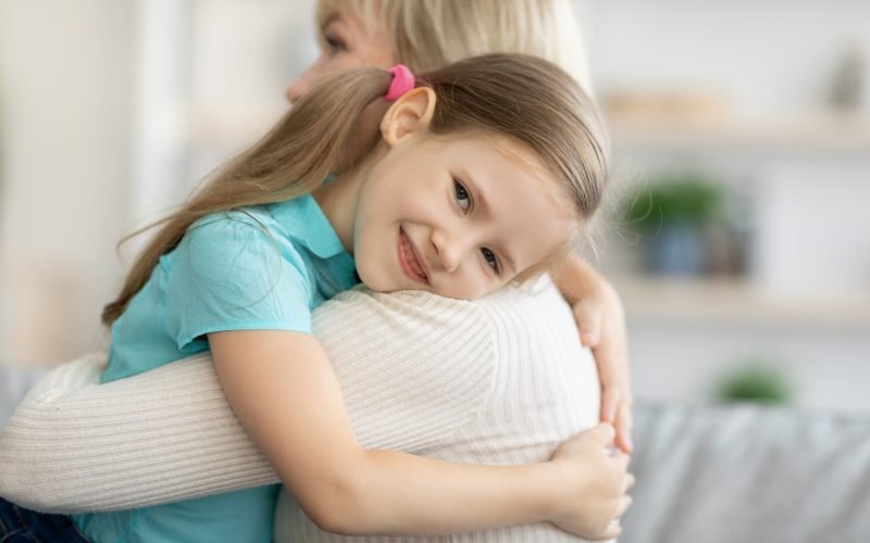 Senior woman hugging child at home. Happy family enjoying kindness, support, care together in living room. Cute girl visiting grandmother, spending time together. Lifestyle moments